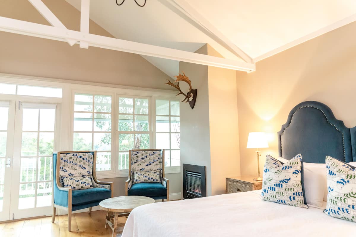A bedroom with a bed, two chairs, a table, a fireplace, and a large window.