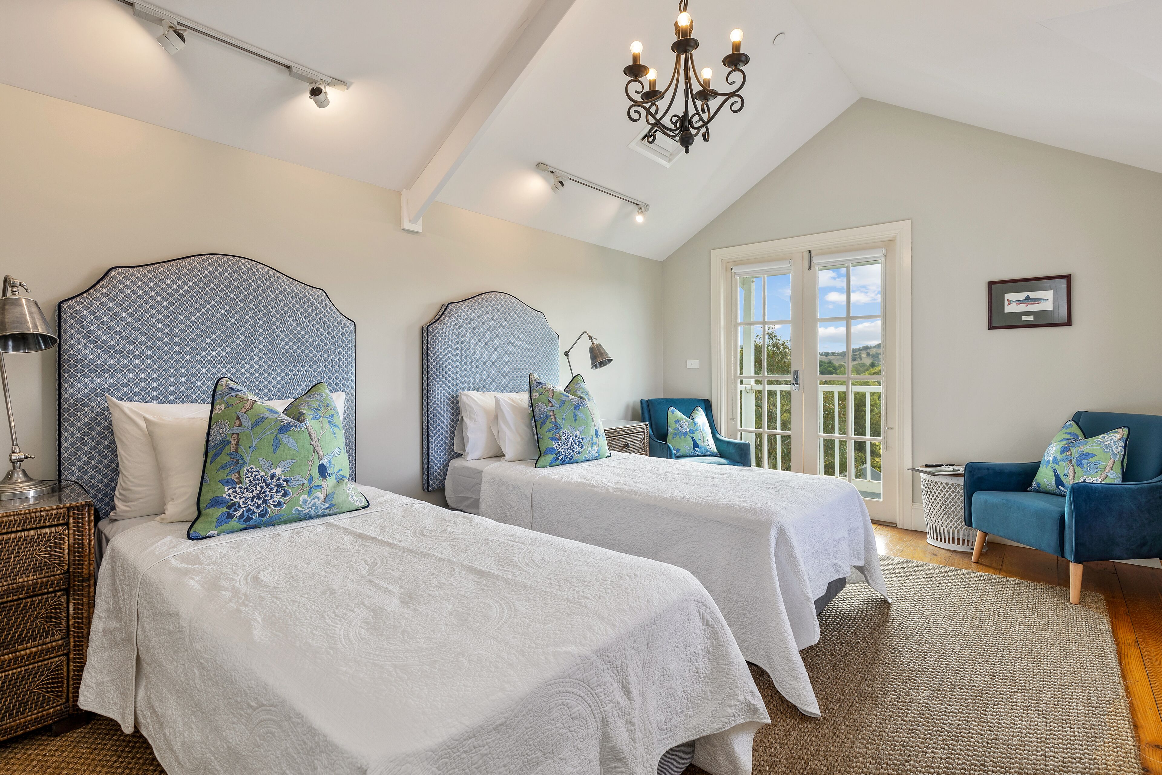 A bedroom with two beds, a chandelier, a blue chair, and a view through a large window.