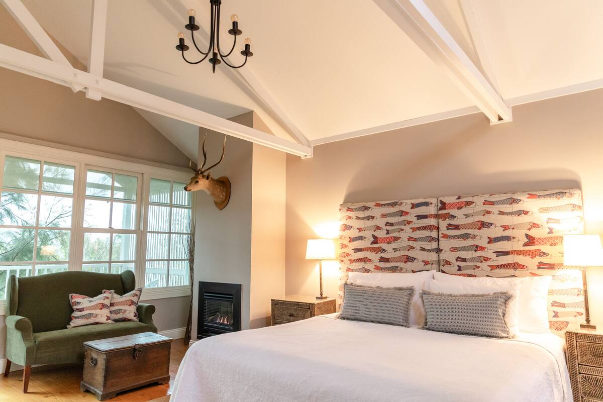 A bedroom with a large bed, a green armchair, a fireplace, and a deer head on the wall.