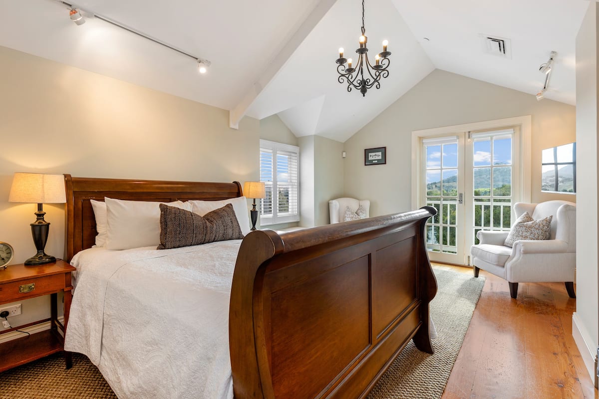 A bedroom with a large bed, a chandelier, a white armchair, and a view of the outdoors through a window.