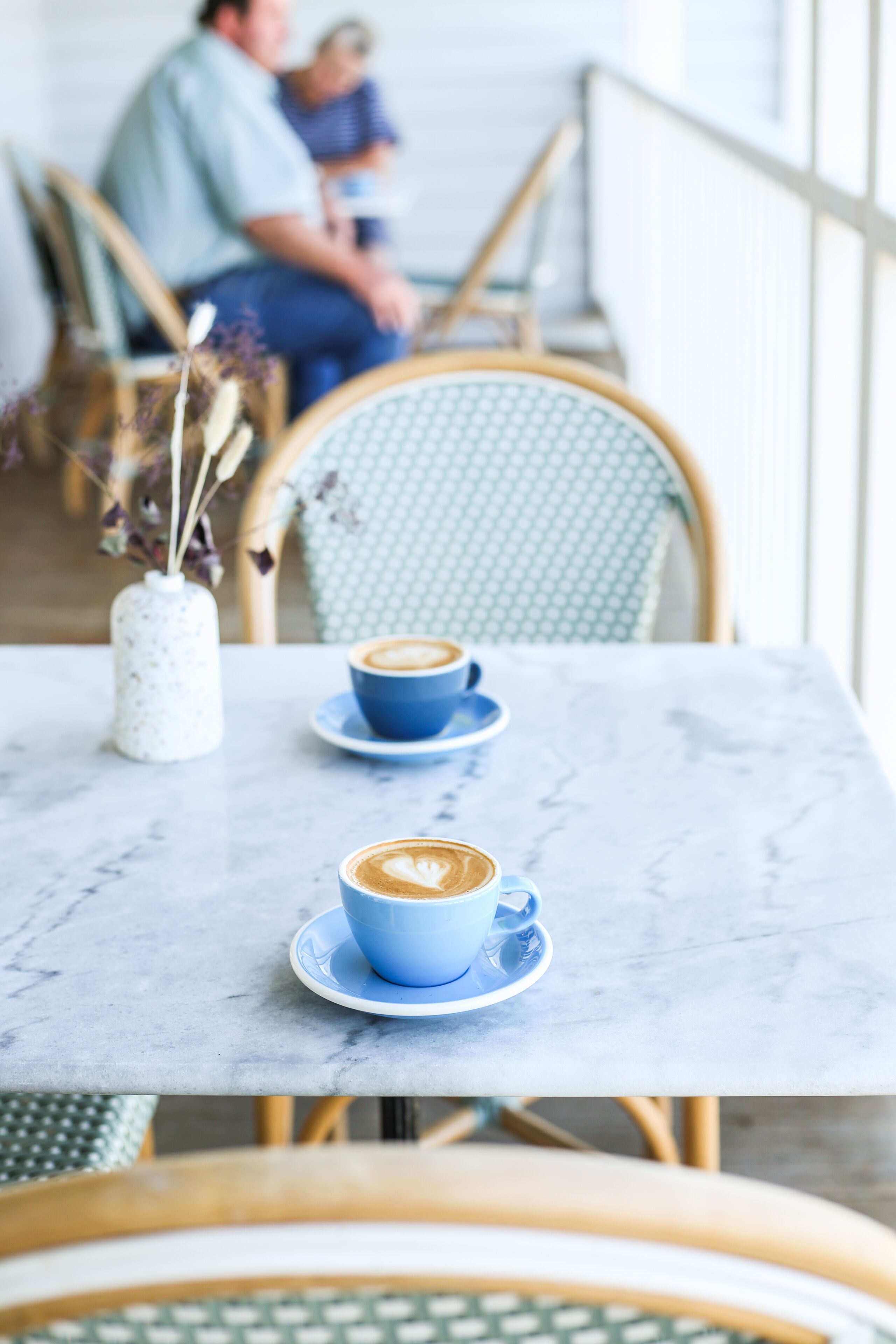 Um café com duas chávenas de café azuis numa mesa de mármore, um vaso branco com flores secas e cadeiras de vime.