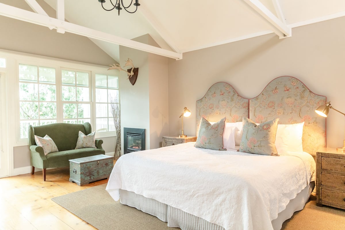 A bedroom with a large bed, a green armchair, a chest of drawers, and a window with a view of greenery.