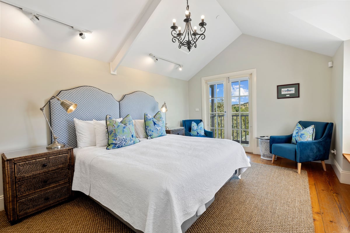A bedroom with a large bed, a chandelier, a blue chair, and a window with a view.