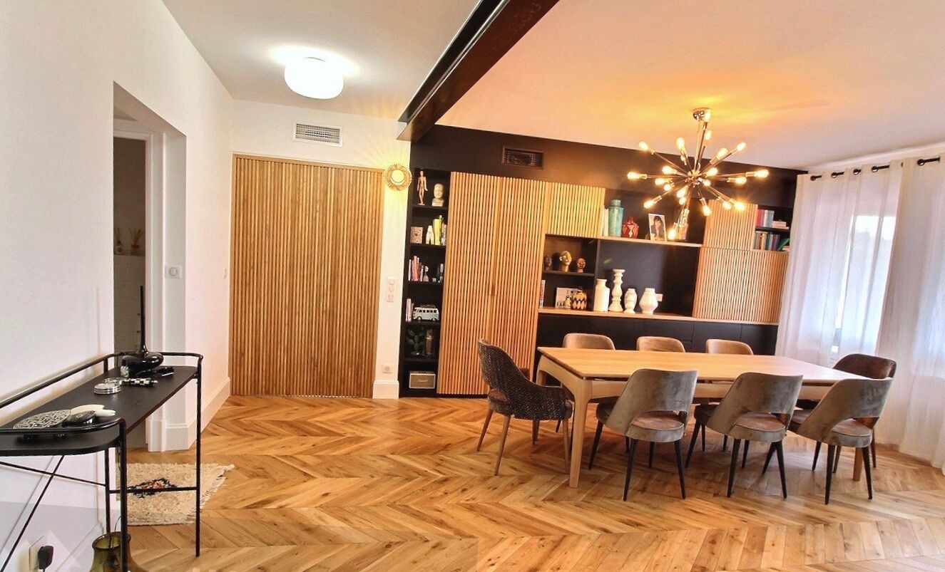Un espace repas doté d’un parquet en bois disposé en chevrons, d’une table centrale entourée de chaises, d’une bibliothèque noire et d’un luminaire décoratif au plafond.