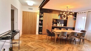 En matplats med ett trägolv i fiskbensmönster, ett stort bord omgivet av stolar, en svart bokhylla och en dekorativ taklampa.