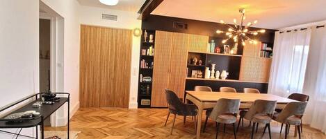 Un espace repas doté d’un parquet en bois disposé en chevrons, d’une table centrale entourée de chaises, d’une bibliothèque noire et d’un luminaire décoratif au plafond.