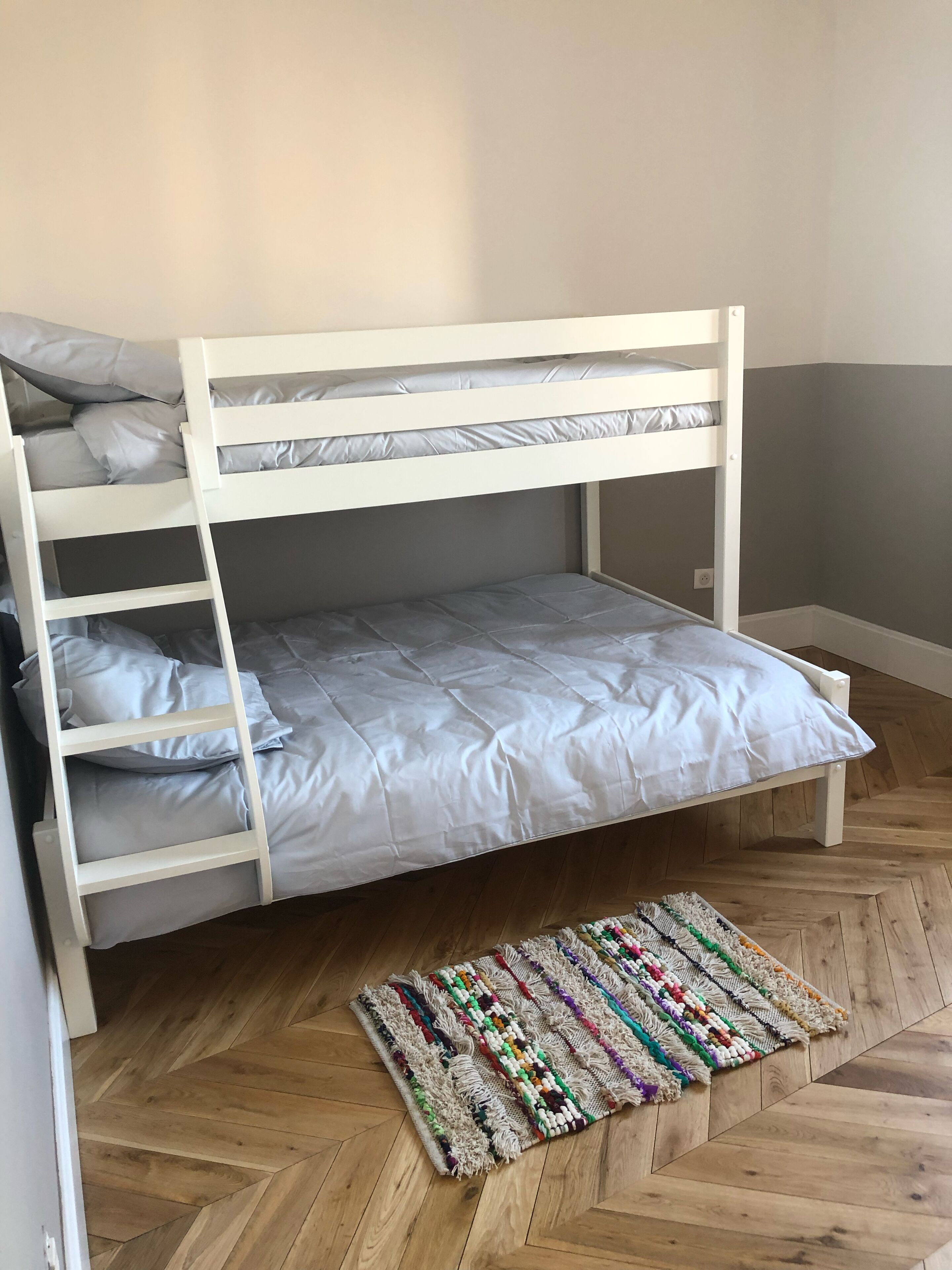A bunk bed with grey bedding, a herringbone wooden floor, and a colorful woven rug.