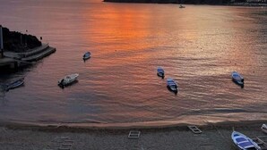 Una playa con barcos anclados en aguas tranquilas, un muelle a la izquierda y una puesta de sol al fondo.