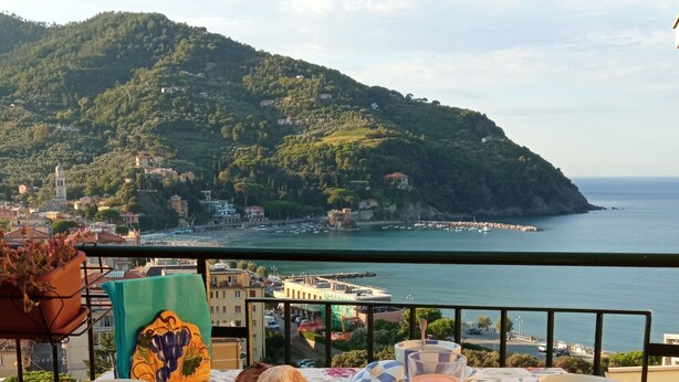 Una mesa servida con alimentos para el desayuno, incluyendo pasteles y jugo, con vistas a un paisaje costero con colinas y el mar.