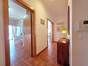 A hallway with a wooden door, a tiled floor, a desk with a lamp, and framed pictures on the wall.