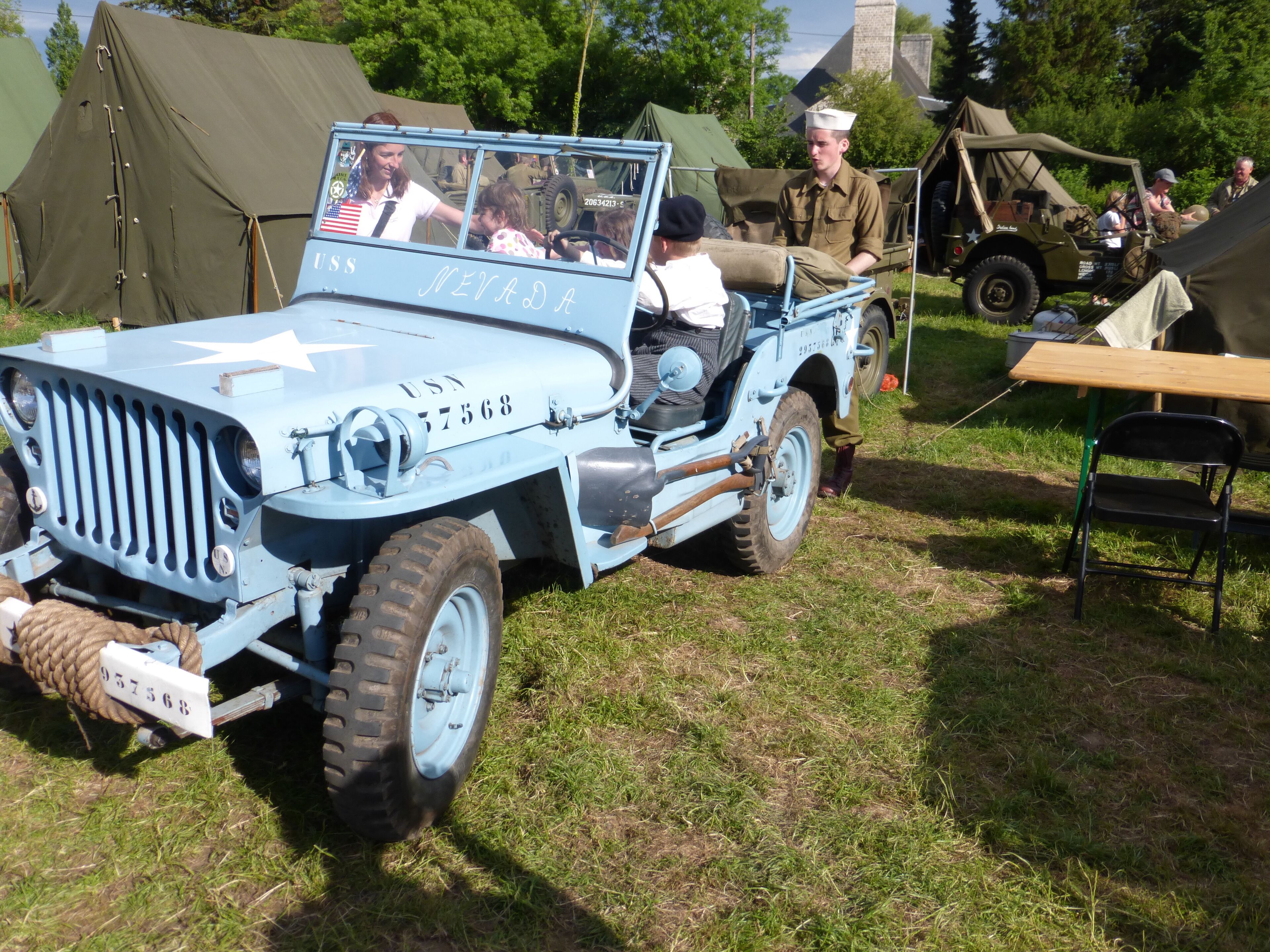 A vintage military jeep with a star emblem and the text "USS Nevada" on the side, parked on grass with tents in the background.