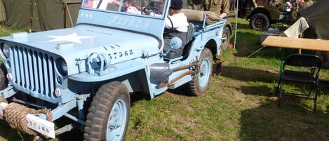 A vintage military jeep with a star emblem and the text "USS Nevada" on the side, parked on grass with tents in the background.