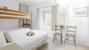 A bedroom with a bunk bed, a wooden chair, a bedside table with a tray of flowers, and a window with curtains. - Lobster Pot - Two Bedroom Cottage, Sleeps 5 (Portloe)