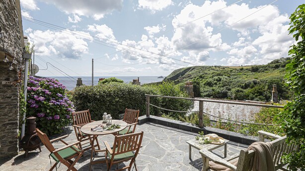 An outdoor patio with a round table and chairs, overlooking a coastal landscape with hills and the sea. - Lobster Pot - Two Bedroom Cottage, Sleeps 5 (Portloe)
