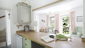 A kitchen with a wooden countertop, a dining area with a chair, and a view of the garden through a window. - Lobster Pot - Two Bedroom Cottage, Sleeps 5 (Portloe)