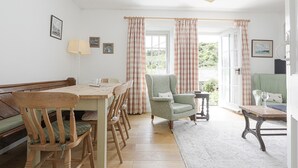 A cozy room with a wooden dining table, chairs, a green armchair, and a small table with a lamp. There are curtains and a view of greenery outside. - Lobster Pot - Two Bedroom Cottage, Sleeps 5 (Portloe)