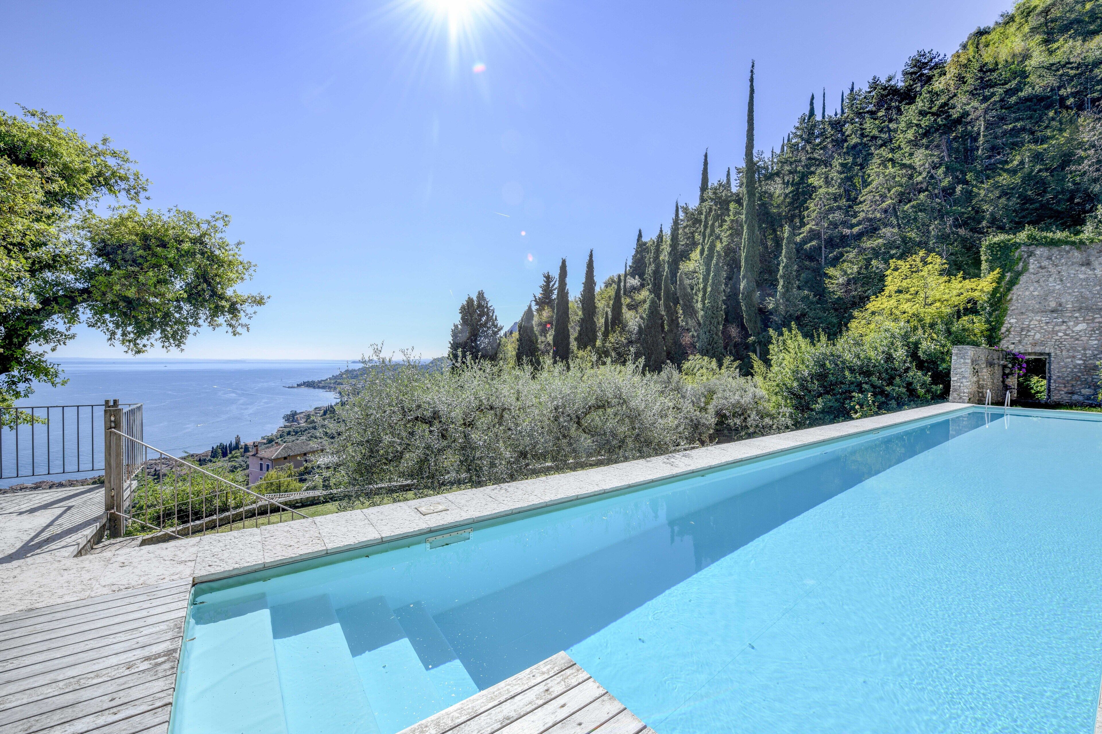 A swimming pool with a wooden deck, surrounded by greenery and overlooking a scenic view of the sea.
