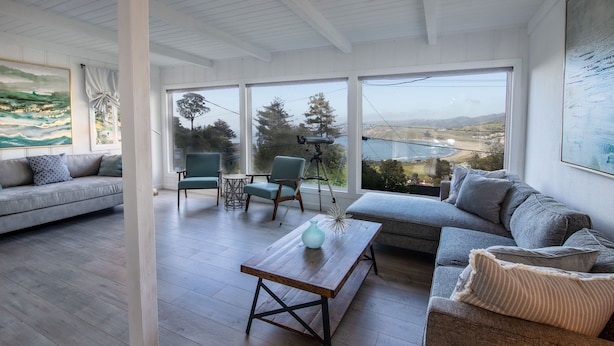 Un salon moderne avec un canapé, une table basse et un fauteuil. Une grande fenêtre offre une vue sur les arbres et les collines.
