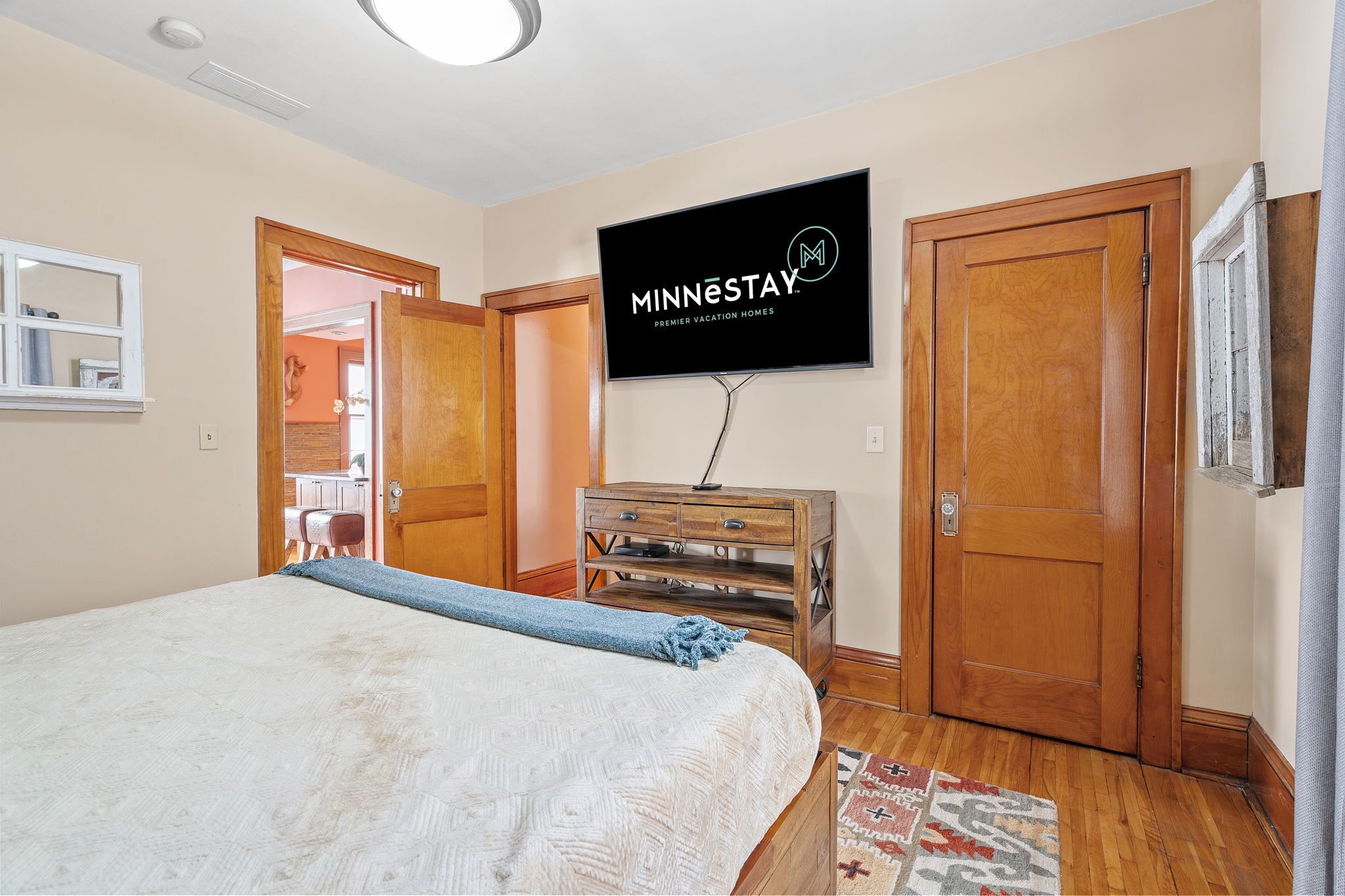 A bedroom with a bed, a TV mounted on the wall, a wooden dresser, and a door leading to another room.