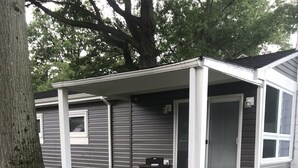 Ein eingeschossiges Mobilheim mit überdachter Veranda, einer kleinen Terrasse mit Tisch und Stühlen sowie einer Moskitotür.