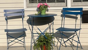 A small outdoor seating area with a round table and two blue chairs, a potted plant, and a window with blinds.