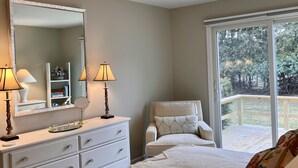 A bedroom with a large bed, a dresser, a mirror, two lamps, a ceiling fan, and a window with a view of trees.