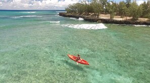 A kayaker in a red kayak on clear turquoise water near a rocky shoreline with sparse vegetation.