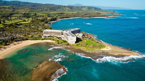 Eine Luftaufnahme eines Küstenresorts mit einem großen Gebäude, direkt am Strand und klarem, blauem Wasser.