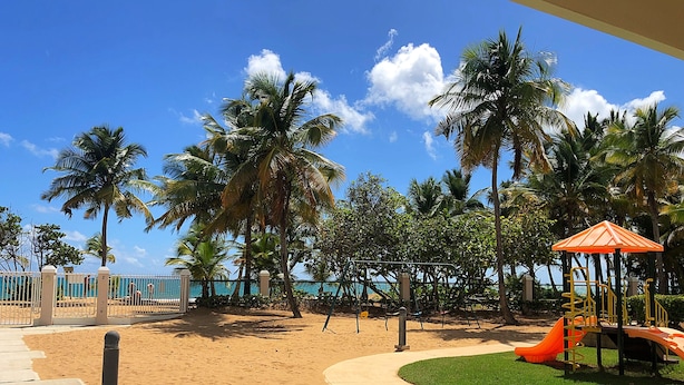 A playground with a slide and climbing structures, surrounded by palm trees and a sandy area. - Beach Oasis - Ground Floor Beach Apartment Steps to the Pool! (Río Grande)