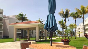 A resort patio with a round wooden table and chairs, a blue umbrella, and palm trees. - Beach Oasis - Ground Floor Beach Apartment Steps to the Pool! (Río Grande)