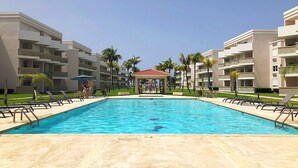A swimming pool surrounded by modern buildings with balconies, palm trees, and a gazebo in the center. - Beach Oasis - Ground Floor Beach Apartment Steps to the Pool! (Río Grande)
