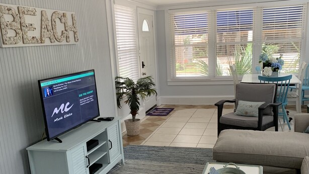 Ein Wohnzimmer mit Fernseher, Deckenventilator, Essbereich mit blauen Stühlen und einer Wand mit dem Schriftzug 'BEACH'.