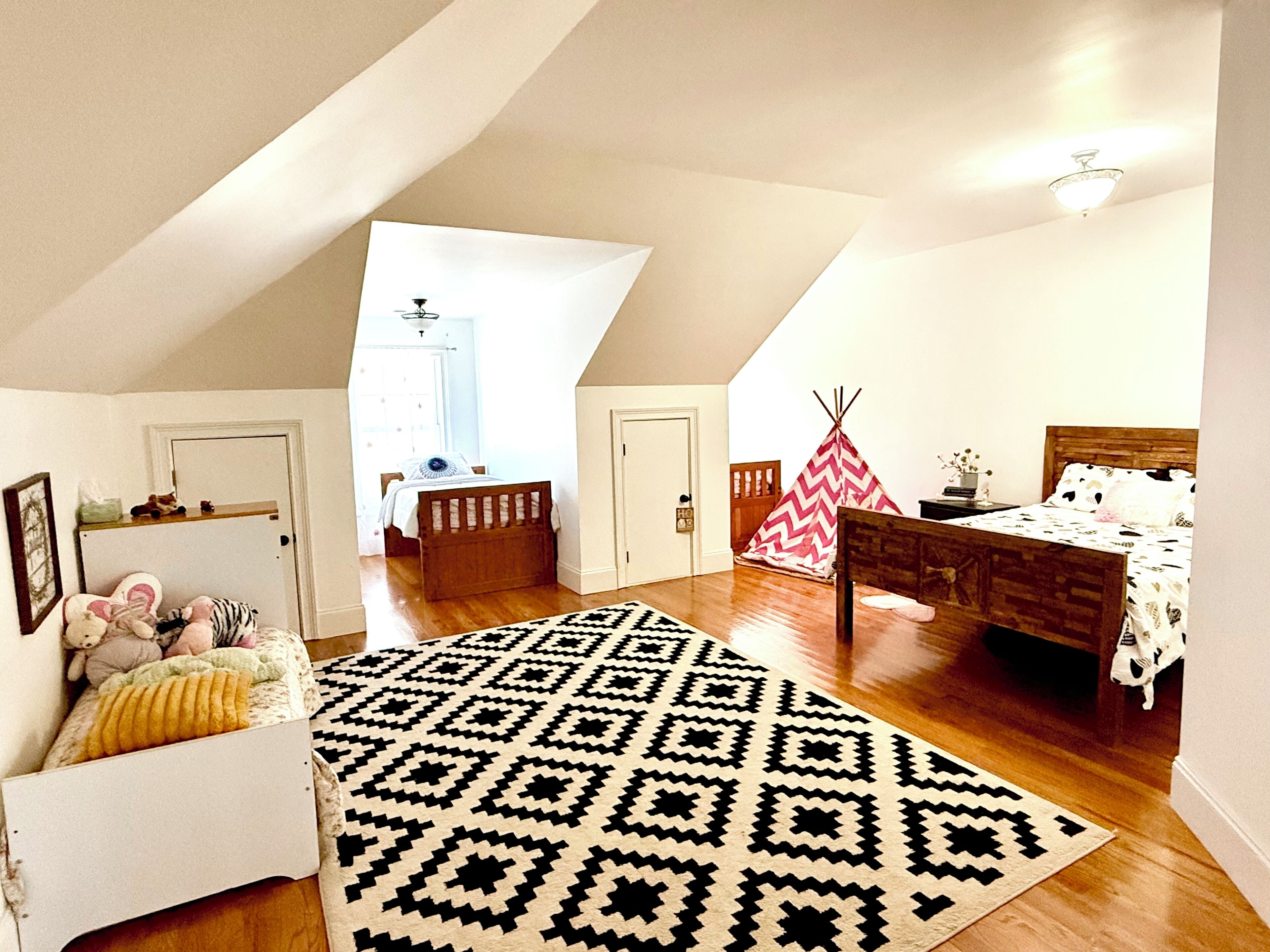 A spacious attic bedroom with a high ceiling, a large window, a wooden bed, a patterned rug, and a teepee decoration.