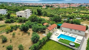 Un quartier résidentiel doté d’une grande piscine, d’un bâtiment blanc avec un toit rouge et offrant une vue sur une ville côtière en arrière-plan.