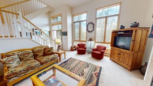 A spacious living room with a wooden staircase, a large window, a sofa, a red armchair, a television, and a coffee table. - Big End Unit, Deck! Near Lake Trail. Easy Drive to Dining, Activities, Events (Frisco)