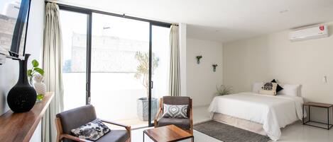 A modern bedroom with a large bed, a sitting area with a chair and a small table, a window with curtains, and a wall-mounted air conditioner.