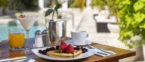 Une table dressée pour le petit-déjeuner avec des gaufres, des fruits rouges, un verre de jus d’orange et une tasse blanche posée sur une table en bois.