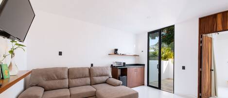 A modern living room with a sectional sofa, a flat-screen TV, a coffee machine, and a sliding glass door leading to a balcony.