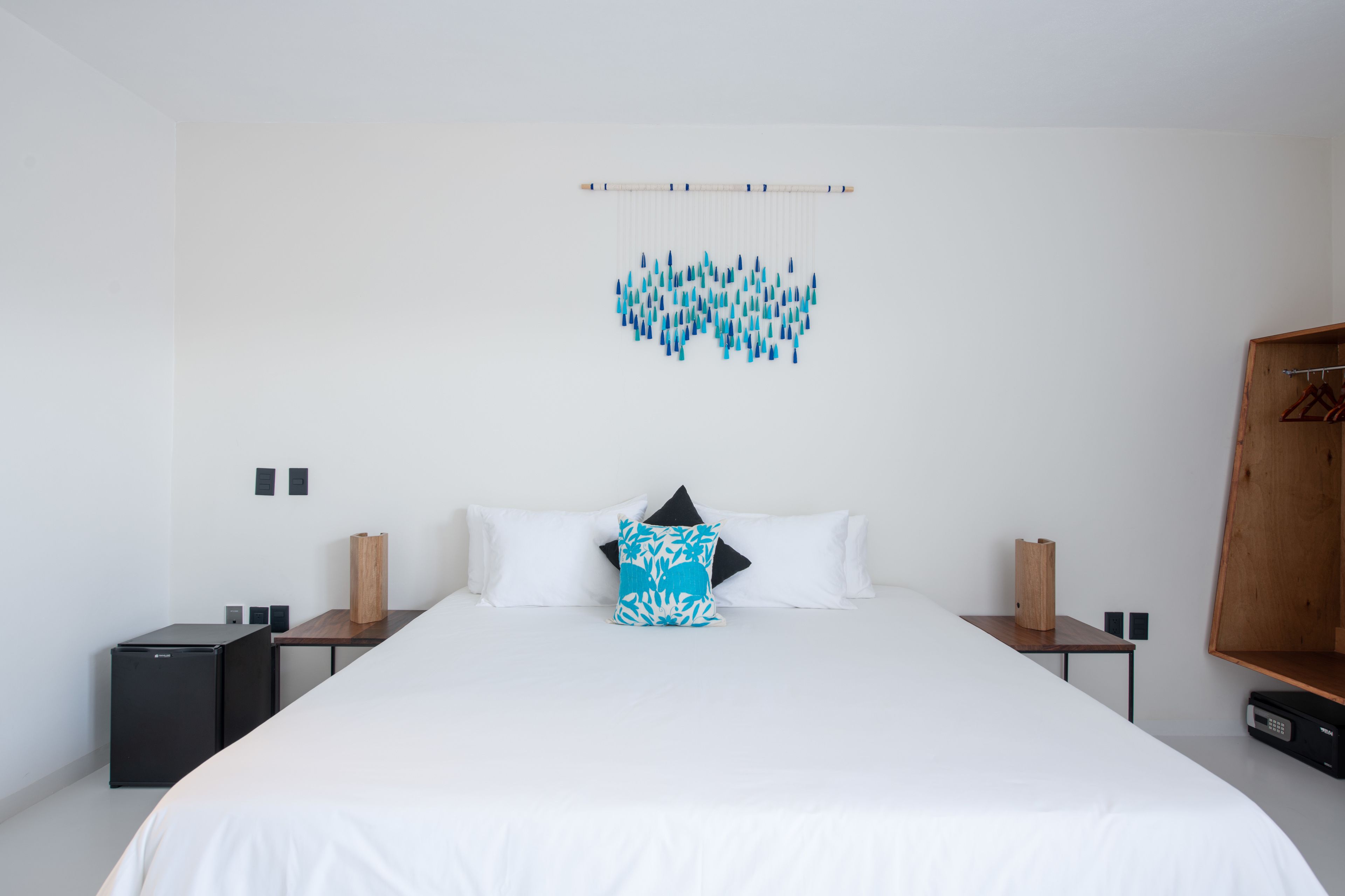 Una habitación de hotel minimalista con una cama grande, dos mesitas de noche, un armario y una obra de arte en la pared.