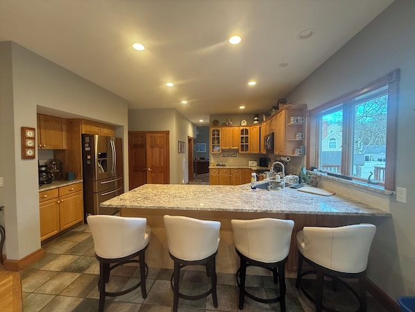 A kitchen with granite countertops, wooden cabinets, a stainless steel refrigerator, and a large island with bar stools. - PPP Peaceful, private with a pool!
House on a Hill in a quiet neighborhood. (Barnstable)