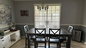 Een eetkamer met een houten tafel en stoelen, een kroonluchter, een raam met zonwering en een kast met keukengerei.