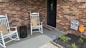 Een bakstenen muur met een zwarte deur, twee witte schommelstoelen, een kleine vuilnisbak, een hangende lamp en een bord met de tekst 'Welkom' naar het terras.