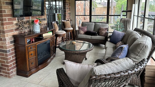 Een zonnig terras met een bakstenen muur, een flatscreen televisie, rieten meubels en een glazen tafel.