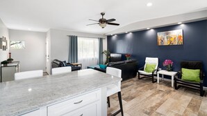 Una sala moderna con una pared de acento azul oscuro, un ventilador de techo, un comedor con encimera de mármol y una zona de estar con un sofá de cuero negro y dos sillones.