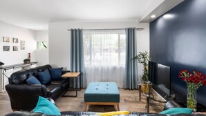 Una sala de estar moderna con un sofá de cuero negro, un reposapiés verde azulado, una mesa de centro de madera, un televisor de pantalla plana y un ventanal con cortinas translúcidas.