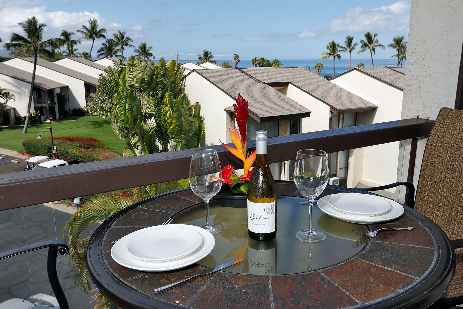A dining table with a bottle of wine, two glasses, and cutlery, set against a balcony with a view of palm trees and a building.