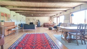 Ein geräumiges Wohnzimmer mit einer großen Holzbalkendecke, einem roten Teppich mit Muster, einem Klavier, Sofas und einem Esstisch mit Stühlen.