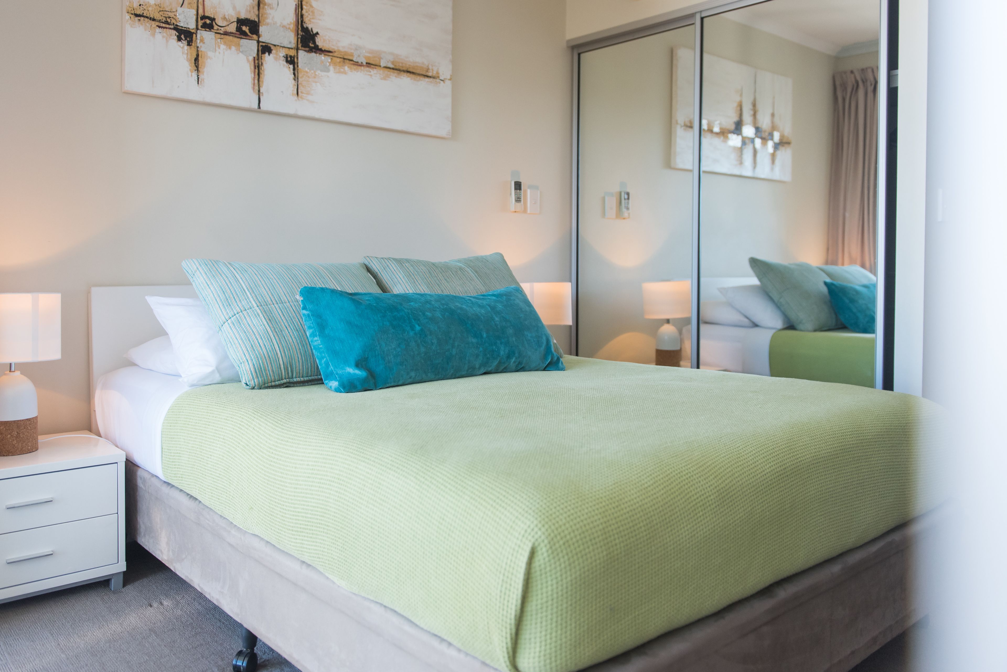 A neatly made bed with a green blanket, a nightstand with a lamp, and a mirror reflecting the room.
