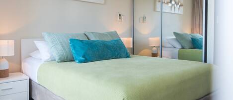 A neatly made bed with a green blanket, a nightstand with a lamp, and a mirror reflecting the room.