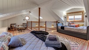 Une chambre spacieuse aménagée en loft, avec un plafond haut, une grande fenêtre dotée de stores, un coin salon confortable avec une table et des chaises, et un espace nuit séparé avec un lit et un canapé.